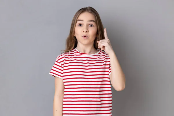 天才的なアイデアで指を指して縞模様のTシャツを着て興奮した少女の肖像画は 突然スマートソリューションを発明したことに驚いている 灰色の背景に隔離された屋内スタジオショット — ストック写真