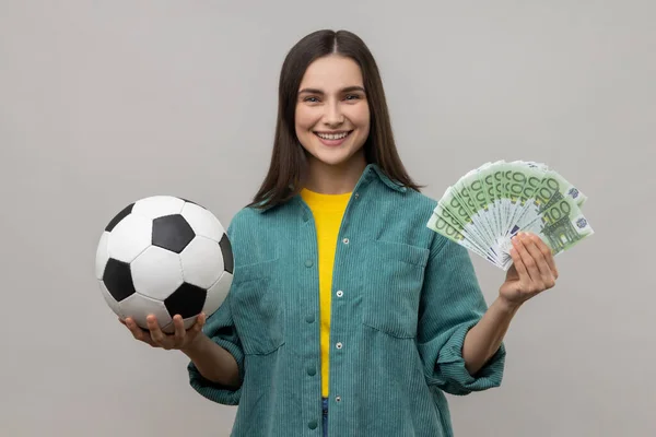 サッカーボールと100ユーロ紙幣の楽しさを示す満足女性の肖像 スポーツのための賭けの多くを獲得し カジュアルなスタイルのジャケットを身に着けている 灰色の背景に隔離された屋内スタジオショット — ストック写真