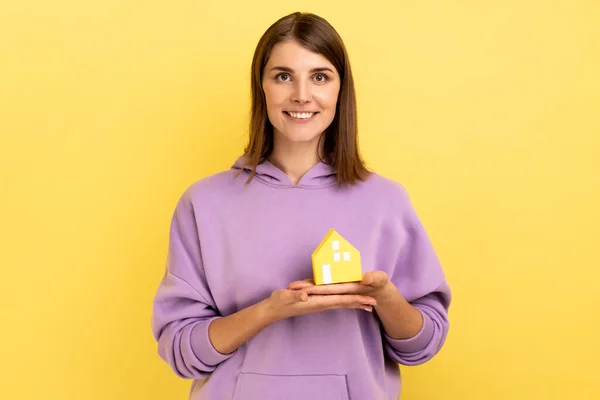 手に小さな紙の家のカメラを見て保持笑顔の女性の肖像画は 彼女自身のアパートについては 紫色のパーカーを着て夢を見る 黄色の背景に隔離された屋内スタジオショット — ストック写真