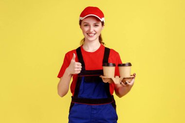 İyimser pozitif kurye kadın portresi kahve fincanlarının önünde duruyor, başparmağını kaldırıyor, tulum giyiyor ve kırmızı şapka takıyor. Sarı arka planda kapalı stüdyo çekimleri.