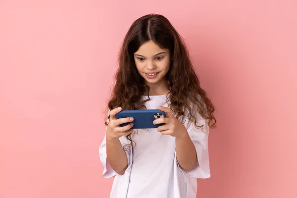 携帯電話でビデオゲームをプレイし ショックを受けた肯定的な表現と携帯電話を使用して白いTシャツを着て驚きの少女の肖像画 ピンクの背景に隔離された屋内スタジオショット — ストック写真