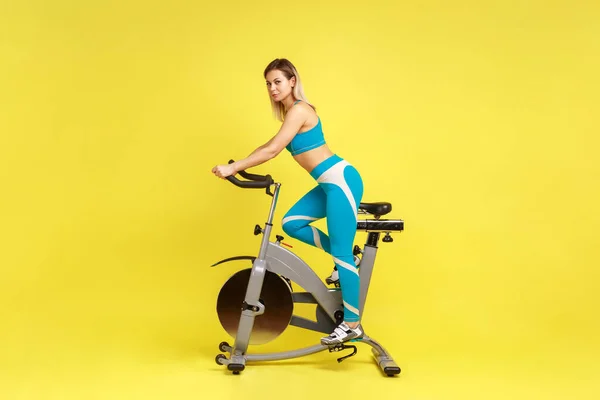 サイドビューブルーのスポーツウェアを着て ブロンドの美しい女性が運動自転車に乗って 健康的なライフスタイルと動機の完全な長さ 黄色の背景に隔離された屋内スタジオショット — ストック写真