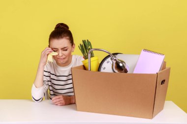 Depresyondaki genç kadın ofis çalışanı elinde karton kutusuyla işyerinde oturuyor ve ağlıyor, kovuluyor. Sarı arka planda kapalı stüdyo çekimleri.