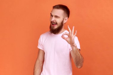 Parmağını kaldırmış sakallı bir adamın portresi, jest gibi, tasvip gösteriyor ve önerilere katılıyor, pembe tişört giyiyor. Turuncu arka planda kapalı stüdyo çekimleri.