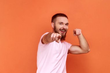 Kamerayı işaret eden, gülümseyen, mutlu seçimler yapan yakışıklı sakallı adamın portresi, pembe tişört giymene ihtiyacımız var. Turuncu arka planda kapalı stüdyo çekimleri.