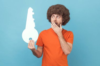 Portrait of shocked man with Afro hairstyle wearing orange T-shirt holding paper key and covering mouth, being surprised, unexpected solution. Indoor studio shot isolated on blue background.