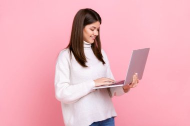 Dizüstü bilgisayarda güzel bir ifadeyle çalışan kadın portresi, en sevdiği serbest çalışma, günlük beyaz süveter giymesi. Pembe arka planda kapalı stüdyo çekimleri.