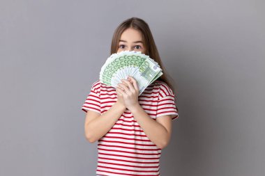 Yüzünün yarısını saklayan çizgili tişört giyen sevimli küçük bir kızın portresi. Yüz avroluk banknotların arkasına saklanıyor, para kokusunun tadını çıkarıyor. Gri arka planda izole edilmiş kapalı stüdyo görüntüleri.
