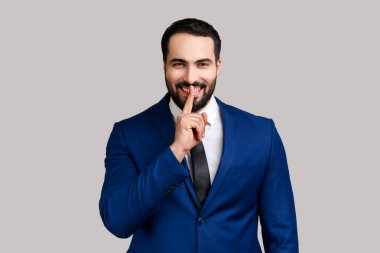Gülümseyen, sakallı, dudak kenarında parmağını tutan, sırlarını saklayan, sürpriz hazırlayan, resmi tarz takım elbiseli olumlu sakallı bir adam. Gri arka planda izole edilmiş kapalı stüdyo görüntüleri.