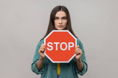 Koyu renk saçlı, elinde kırmızı dur işareti olan ciddi bir kadın, kameraya negatif agresif bir ifadeyle bakıyor, yasağı gösteriyor, günlük tarz ceket giyiyor. Gri arka planda izole edilmiş kapalı stüdyo görüntüleri.