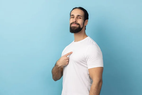 Portrait of self confident man with beard wearing white T-shirt pointing at himself, feeling proud and self-important, having big ego. Indoor studio shot isolated on blue background.