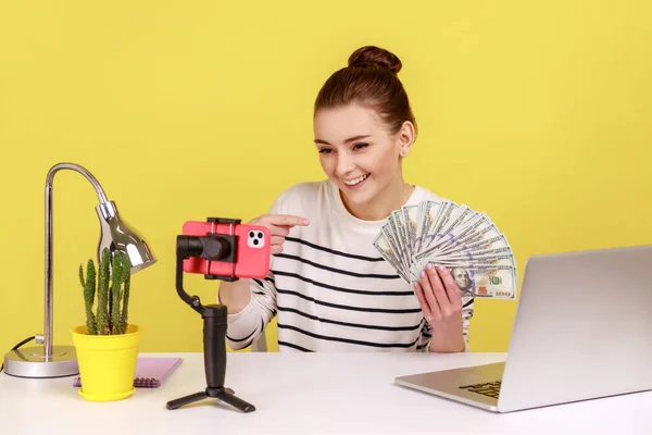Woman pointing finger at dollar cash, recording video to her followers, talking with subscribers, boasting salary raising, sitting at workplace. Indoor studio studio shot isolated on yellow background