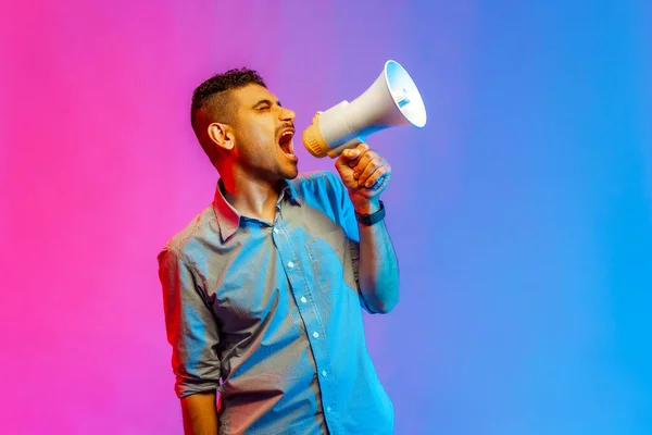 Portrait of aggressive man in shirt screaming at megaphone, making announce, protesting, looking away with angry expression. Indoor studio shot isolated on colorful neon light background.