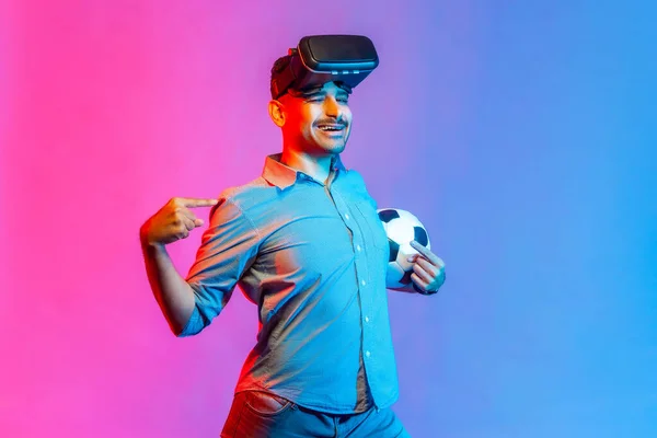 Happy smiling man in shirt wearing virtual reality glasses, standing with satisfied expression, pointing at himself with proud. Indoor studio shot isolated on colorful neon light background.