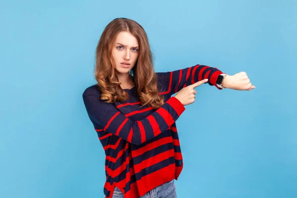 Time is gone! Young serious woman wearing striped casual style sweater showing time on his watch, looking at camera with bossy expression. Indoor studio shot isolated on blue background.