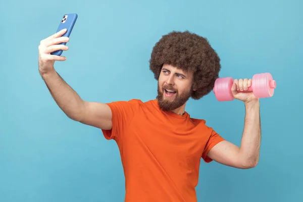 Positive man with Afro hairstyle wearing orange T-shirt making video call and raised arm with dumbbell, taking selfie or broadcasting livestream. Indoor studio shot isolated on blue background.