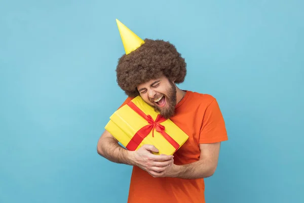Portrait of satisfied man with Afro hairstyle wearing orange T-shirt and party cone embracing yellow gift box, being happy to get present. Indoor studio shot isolated on blue background.