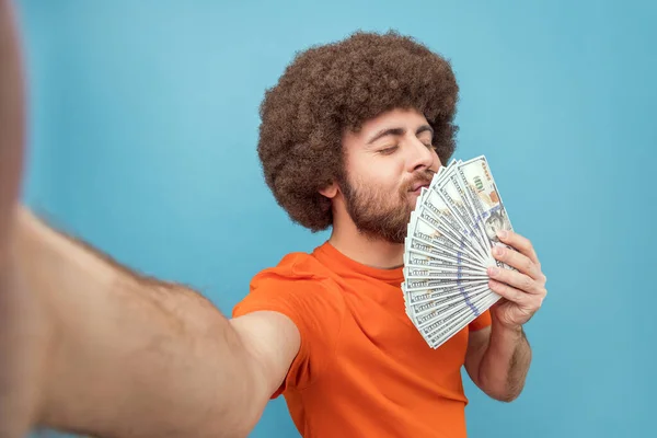 Portrait of satisfied rich man with Afro hairstyle wearing orange T-shirt holding and smelling dollar banknotes, being delighted to get salary, POV. Indoor studio shot isolated on blue background.