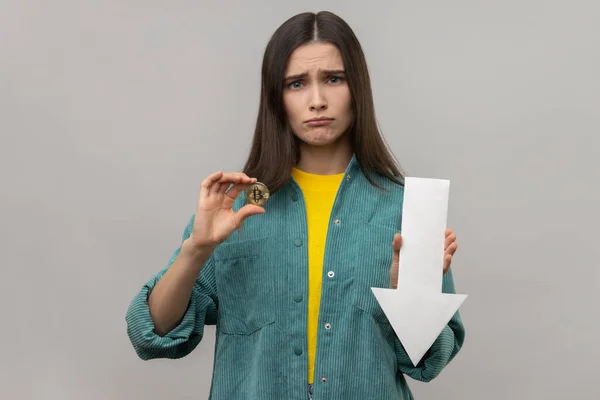 Portrait of young adult unhappy woman showing downgrade of bitcoin, expressing sad upset emotions, looking at camera, wearing casual style jacket. Indoor studio shot isolated on gray background.