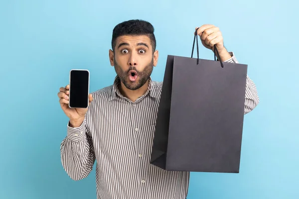 Amazed businessman with beard holding white display smartphone and black paper bag, online shopping, fast delivery, marketplace, wearing striped shirt. Indoor studio shot isolated on blue background.