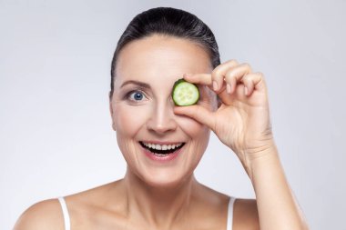 Skin care natural beauty and cosmetology concept. Closeup of excited middle aged woman covering eye with slice of fresh cucumber, makes mask for face. Indoor studio shot isolated on gray background.
