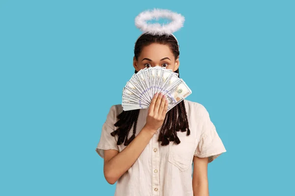 Angelic happy rich woman with black dreadlocks and nimbus on head peeking out of dollar banknotes, rejoicing big profit, wearing white shirt. Indoor studio shot isolated on blue background.