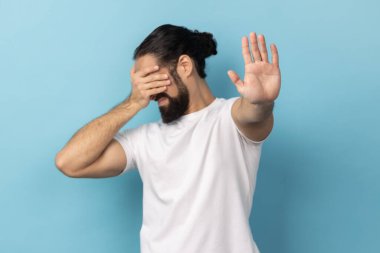 Bakmak istemiyorum. Beyaz tişörtlü, sakallı bir adam gözlerini üzerinden ayırmıyor ve utanç verici bir şeyi izlemekten kaçınıyor, yasaklama hareketi gösteriyor. Mavi arka planda kapalı stüdyo çekimi.
