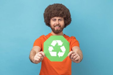 Turuncu tişörtlü Afro saçlı bir adamın portresi elinde atık geri dönüşüm sembolü, ekoloji ve çevre kirliliği hakkında endişeleniyor. Mavi arka planda kapalı stüdyo çekimi.