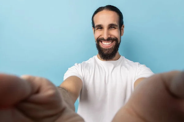 Man blogger with beard in white T-shirt looking at camera with toothy smile, being in good mood, broadcasting livestream, POV, point of view of photo. Indoor studio shot isolated on blue background.