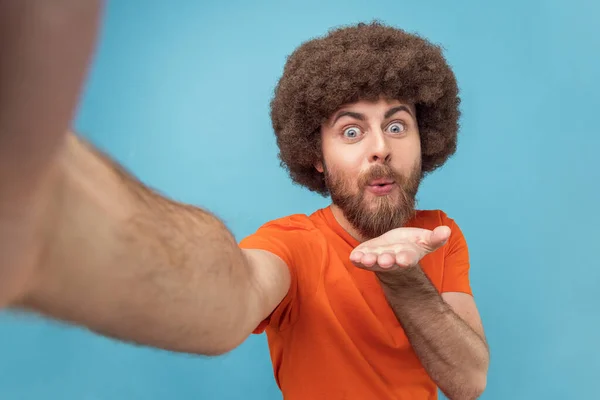 Portrait of handsome man with Afro hairstyle wearing orange T-shirt sending air kisses to his subscribers, posing with pout lips, point of view photo. Indoor studio shot isolated on blue background.