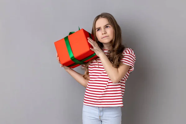 Portrait of charming serious little girl wearing striped T-shirt shaking wrapped present box, being interested what inside, looking away, Indoor studio shot isolated on gray background.