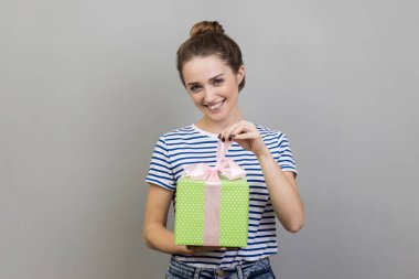 Çizgili tişört giyen bir kadının portresi hediye kutusunu açıyor, bayramı kutlamak için hediye paketini açıyor, kurdele çekiyor, kameraya bakıyor. Gri arka planda izole edilmiş kapalı stüdyo görüntüleri.