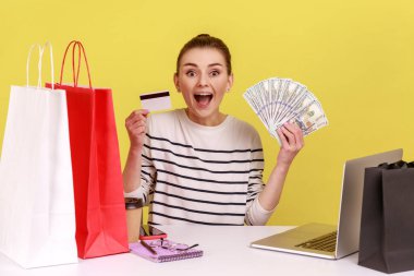 Bank loan, electronic money. Amazed woman sitting office workplace with laptop, holding dollars and credit card, looking at camera with smile. Indoor studio studio shot isolated on yellow background.