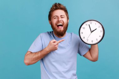 Portrait of happy positive handsome bearded man pointing at wall clock with finger, looking at camera with toothy smile, laughing. Indoor studio shot isolated on blue background.