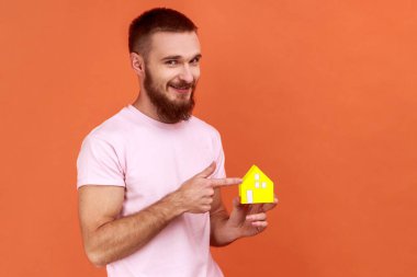 Sakallı bir adamın portresi çizilmiş kalbi, yetimhanesi ya da barınağı olan pembe tişörtü olan küçük kağıt evi işaret ediyor. Turuncu arka planda kapalı stüdyo çekimleri.