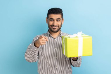 Mutlu sakallı iş adamı hediye kutusu tutuyor ve kamerayı işaret ediyor, seni seçiyor, bonus gösteriyor, tatilde tebrik ediyor, çizgili gömlek giyiyor. Mavi arka planda kapalı stüdyo çekimi