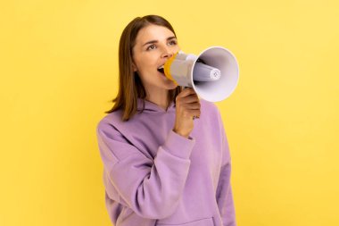 Portrait of beautiful woman screaming loudly in megaphone, announcing important advertisement, protesting, wearing purple hoodie. Indoor studio shot isolated on yellow background.