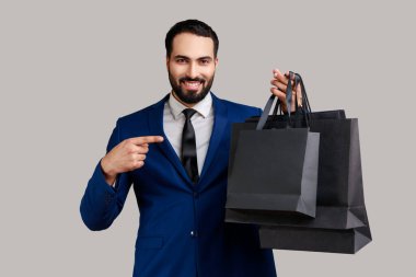 Elindeki kağıt torbaları işaret eden sakallı adam, dişlek gülümseme, alışveriş ve satış ile bakıyor, resmi stil takım elbise giyiyor. Gri arka planda izole edilmiş kapalı stüdyo görüntüleri.