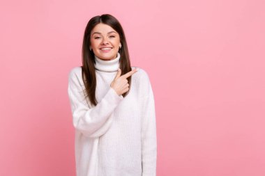 Çekici genç kız reklam içeriği için boş yeri işaret ediyor, ticari ürün için fotokopi alanı, beyaz günlük tarzda süveter giyiyor. Pembe arka planda kapalı stüdyo çekimleri.