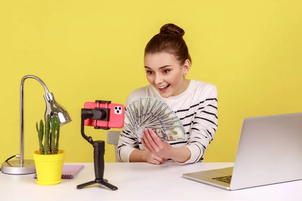 Excited woman holding dollar banknotes, recording video to subscribers, talking with followers, boasting salary raising, sitting at workplace. Indoor studio studio shot isolated on yellow background
