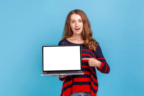 Astonished woman wearing striped casual style sweater, pointing at blank laptop screen with surprised expression, shocked advertisement. Indoor studio shot isolated on blue background.