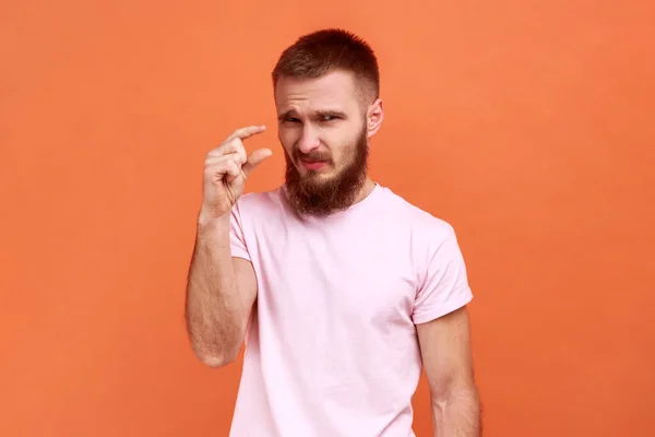 Too small amount. Portrait of bearded man showing a little bit gesture, dissatisfied with low rating, measuring scale, wearing pink T-shirt. Indoor studio shot isolated on orange background.