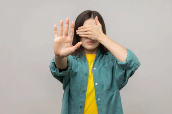 I don't want to watch. Portrait of scared confused adult woman covering eyes, showing stop gesture, afraid and shy to look, wearing casual style jacket. Indoor studio shot isolated on gray background.