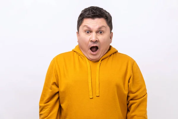 Portrait of excited man with open mouth and big amazed eyes, looking surprised and silly at camera, wondered expression, wearing urban style hoodie. Indoor studio shot isolated on white background.