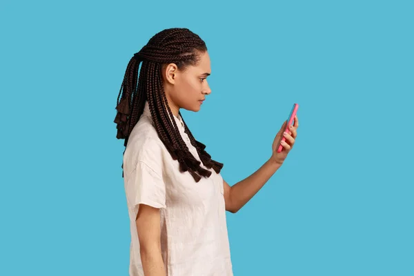 Side view of woman with dreadlocks reads message on modern mobile phone, surfes social media, has concentrated look in display, updates information. Indoor studio shot isolated on blue background.