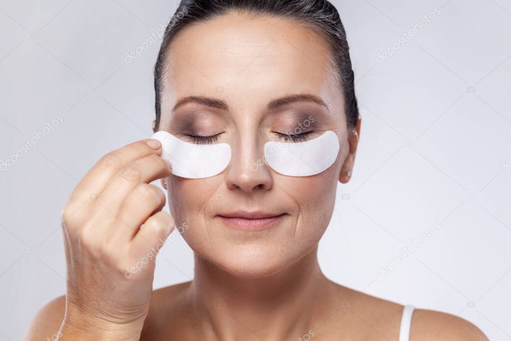 Closeup portrait of middle aged woman with cosmetic collagen patches under eyes, enjoys flawless of skin, has well cared complexion. Indoor studio shot isolated on gray background.