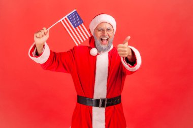 Smiling positive elderly man with gray beard wearing santa claus costume holding USA flag, showing thumb up to camera, having patriotic mood. Indoor studio shot isolated on red background.