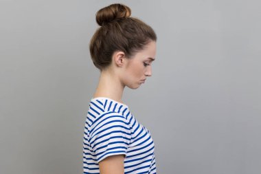 Side view of sad upset young adult woman wearing striped T-shirt expressing sadness, hearing bad news, being in bad mood. Indoor studio shot isolated on gray background.