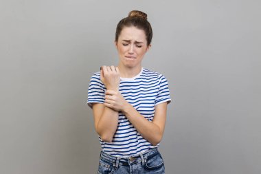 Portrait of unhappy sick woman wearing striped T-shirt touching painful hand, suffering trauma, sprain wrist, feeling ache of carpal tunnel syndrome. Indoor studio shot isolated on gray background.