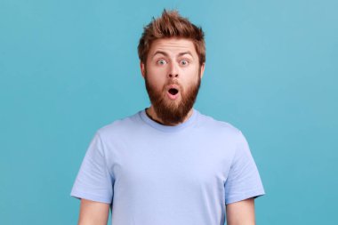 Portrait of shocked bearded man gasps from wonder, holds breath, stares at something horrible keeps mouth opened, being astonished. Indoor studio shot isolated on blue background.